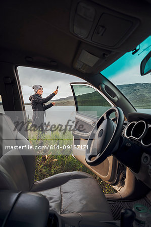 Young woman standing beside Dillon Reservoir, holding smartphone, view through parked car, Silverthorne, Colorado, USA