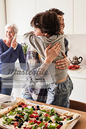 Mother and son hugging in kitchen, grandmother in background using smartphone