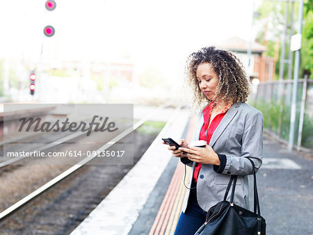 Mid adult woman standing on train platform, using smartphone, holding disposable coffee cup