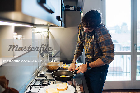 Mid adult man preparing fried eggs at kitchen hob while making smartphone call