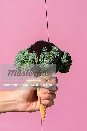 Studio shot of man's hand holding ice cream cone with broccoli on top and pouring chocolate sauce, against pink background