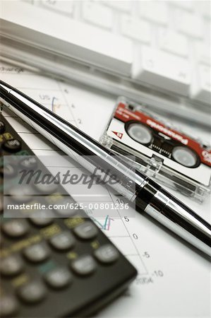 Calculator, computer keyboard, micro cassette recording tape and pen on top of financial graph, close-up