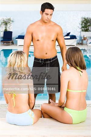 Mid adult man standing in front of two young women at the poolside