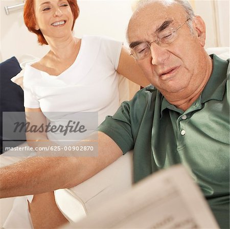 Close-up of a senior man reading a newspaper with a senior woman sitting behind him