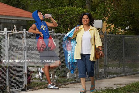 Mature woman walking with a teenage boy leaning on a chain- link fence and drinking water beside her