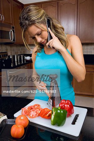 Young woman talking on a mobile phone and grating a carrot in the kitchen