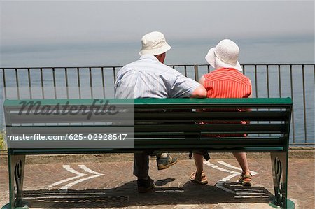 Rear view of a couple sitting on a bench at seaside, Via Aniello Califano, Bay of Naples, Sorrento, Sorrentine Peninsula, Naples Province, Campania, Italy