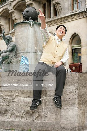 Asian man sits on the brim of a fountain and is laughing while pointing at something