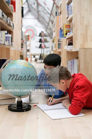 Two boys lying on a floor next to a globe and making notes, fully_released