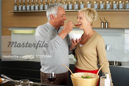 Gray-haired man lets a blond woman taste something he has cooked