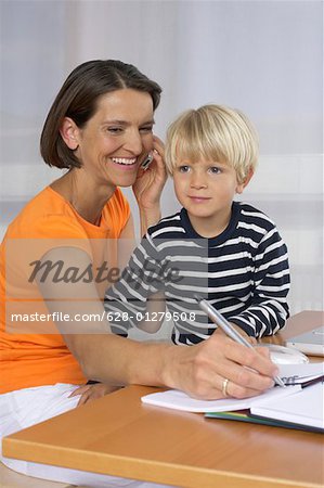 Mother phoning with a mobile phone and taking notes, son (4-5 Years) sitting on her lap