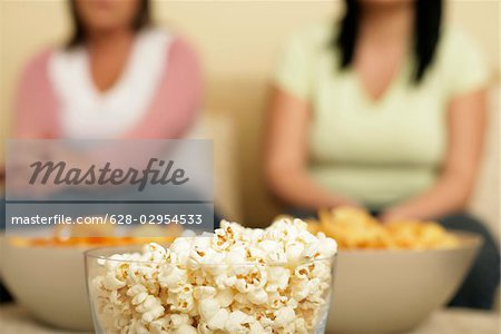 Two full figured women sitting behind three bowls filled with popcorn and crisps, selective focus