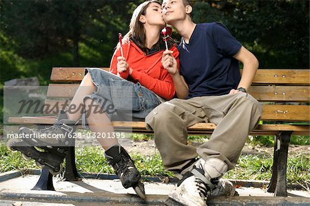 Teenage couple with inline skates sitting on a bench and holding lollypops in their hands, she kissing him