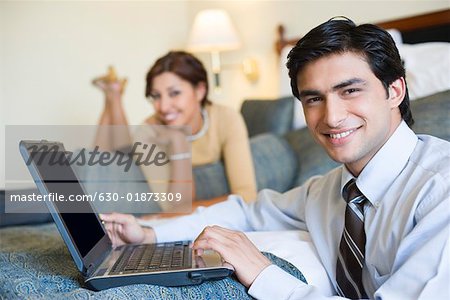 Portrait of a businessman working on a laptop and with a young woman lying on the bed in the background