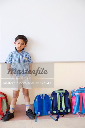 Portrait of a schoolboy standing beside schoolbags in a classroom