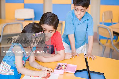 Side profile of a girl drawing on a sheet of paper with two boys looking at her drawing sheet