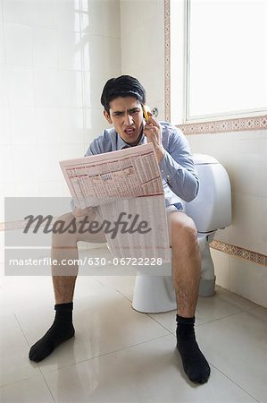 Businessman talking on a mobile phone and reading a newspaper on a toilet seat
