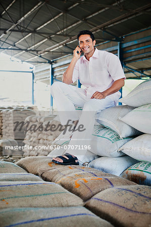 Man sitting on stack of wheat sacks and talking on a mobile phone in a warehouse, Anaj Mandi, Sohna, Gurgaon, Haryana, India Man sitting on stack of wheat sacks and talking on a mobile phone in a warehouse, Anaj Mandi, Sohna, Gurgaon, Haryana, India