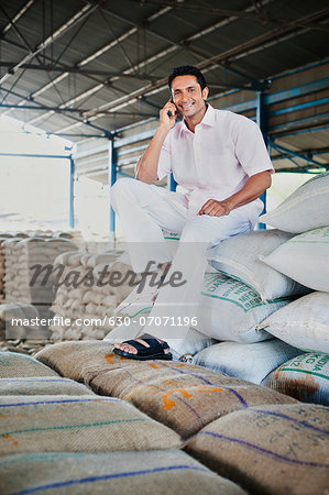 Man sitting on stack of wheat sacks and talking on a mobile phone in a warehouse, Anaj Mandi, Sohna, Gurgaon, Haryana, India