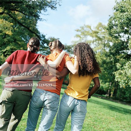 Three young women walking on grass with arms around each other, rear view