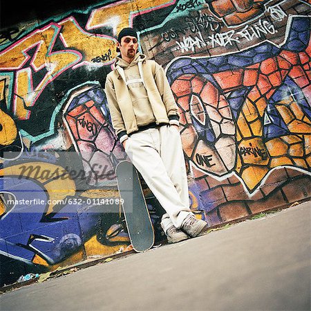 Young man with skateboard leaning against graffitied wall, portrait