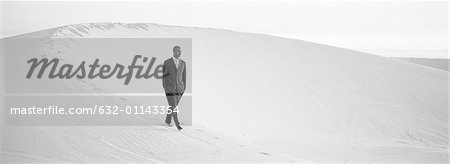 Businessman walking across ridge of dune with hands in pocket, b&w, panoramic view