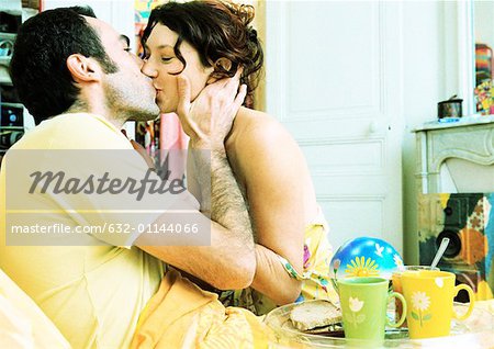 Man in bed with breakfast tray on lap, kissing woman, side view.