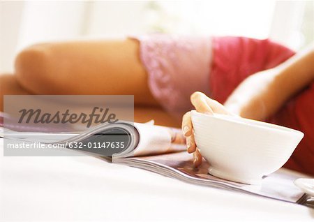 Woman lying on bed, mid-section, blurred, with magazine and bowl in focus in foreground, close-up.