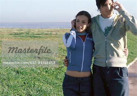 Young couple standing with arms around eachother, both talking on cell phones