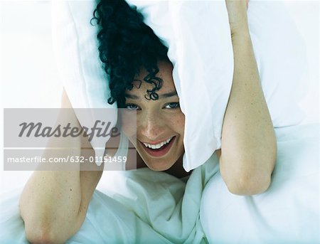 Woman in bed holding pillows to both sides of head, looking at camera