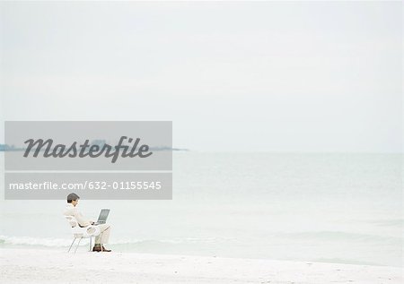 Businessman using laptop on beach, mid-distance