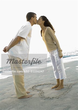Newlyweds, woman standing inside heart drawn in sand, leaning forward to kiss husband