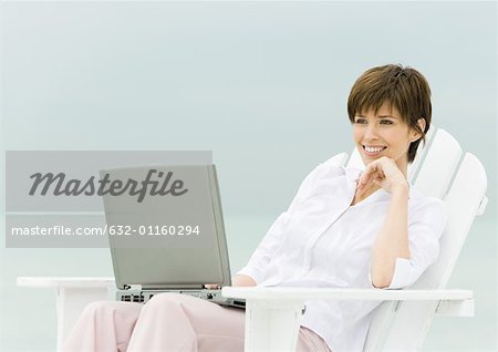 Woman sitting in deckchair using laptop, sea in background