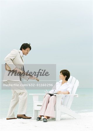 On beach, man in suit serenading woman sitting in deck chair