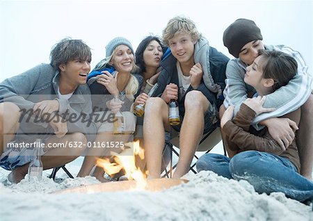 Group of young adults sitting around beach campfire, with bottles of beer