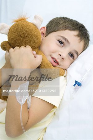 Boy lying in hospital bed with IV drip in arm, hugging stuffed animal