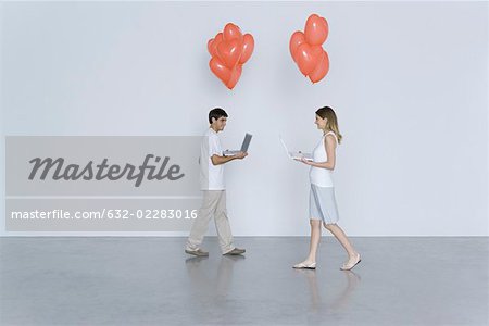 Young man and woman walking toward each other, both carrying laptop computers and heart balloons