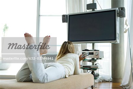 Female lying on stomach watching flat screen TV with surround sound, remote control in hand, rear view