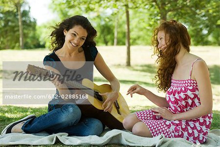 Two young women sitting on blanket outdoors, one playing acoustic guitar