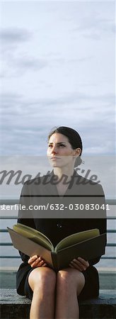 Woman sitting on bench with open book on lap, looking away in thought