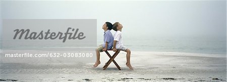 Two boys sitting back to back on chair on beach