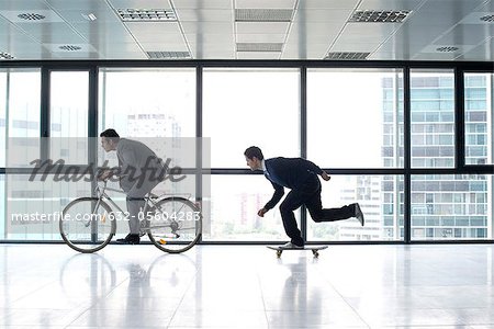 One businessman riding bicycle, the other skateboarding