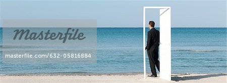 Businessman standing on beach looking at ocean, seen through open door