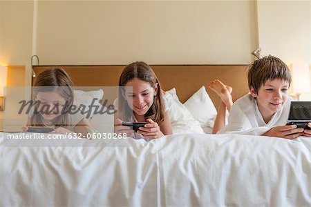 Siblings playing handheld video games on bed