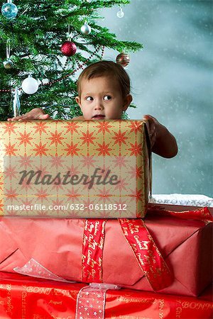 Baby girl standing behind stack of Christmas gifts with mischievous expression