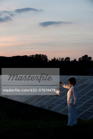 Girl standing in front of solar panels at twilight with old-fashioned lantern in hand