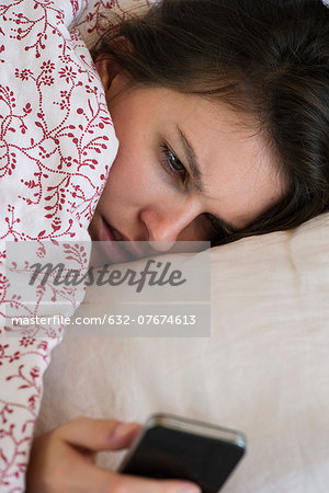 Woman lying bed looking at smartphone with unhappy expression on face Woman lying bed looking at smartphone with unhappy expression on face