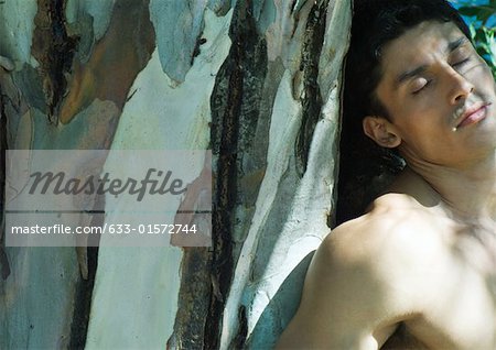 Barechested man leaning against tree trunk