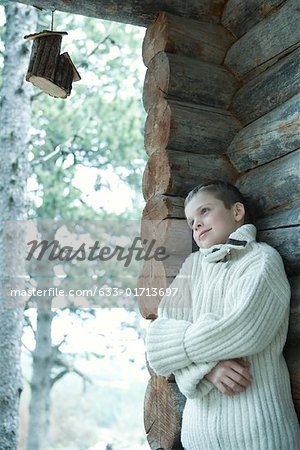 Boy wearing wool sweater, arms folded, leaning against log cabin