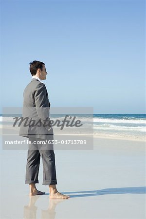 Businessman standing barefoot on beach with hands in pockets, side view